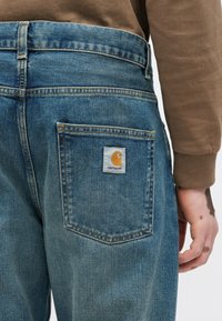 Jasnoniebieskie dżinsy o luźnym kroju, z naszywką z logo na tylnej kieszeni, widocznymi przeszyciami oraz lekko spranym wyglądem.