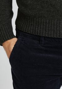 Dunkelgrauer gerippter Pullover, getragen über dunkelblauen Cordhosen. Nahaufnahme einer Hand in der Tasche, die die Textur und Passform der Kleidungsstücke hervorhebt.