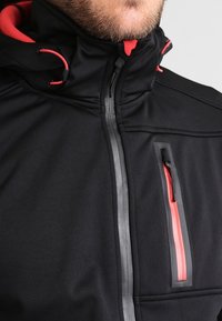 Chaqueta negra con un alto cuello, acentos rojos contrastantes, bolsillo vertical con cremallera y material texturizado, presentando un diseño elegante.