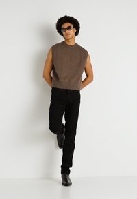 Suéter de punto sin mangas marrón, pantalones de mezclilla negros y botas negras hasta el tobillo. El modelo está de pie con una pierna doblada, usando gafas de sol.