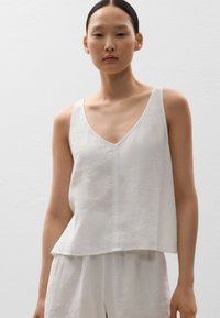 Top din in cu decoare albă, cu decolteu în V adânc și croială lejeră, având o textură subtilă și linii curate. Asortat cu pantaloni ușori, de aceeași nuanță.