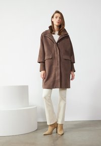 Manteau en mélange de laine marron avec un col haut, fermeture à boutons-pression et poches plaquées. Porté sur un haut blanc, associé à un pantalon beige et des talons.
