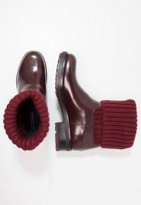 Botas de cuero burdeos con un puño de punto acanalado. Textura suave, punta redondeada y suela de goma gruesa con patrones de agarre.