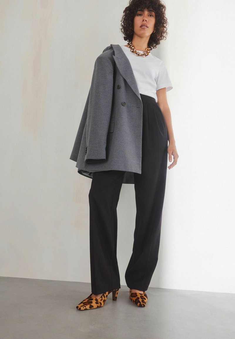 Manteau gris croisé sur un t-shirt blanc, associé à un pantalon noir taille haute et des talons en léopard, avec un collier imposant.