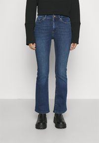 Person som bär mörkblå bootcut-jeans, svart långärmad topp och svarta kängor med tjock sula, står mot en enfärgad bakgrund.