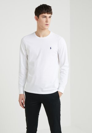 Fehér, hosszú ujjú pamut póló, kerek nyakkal és egy kis sötétkék logóval a mellkason, fekete, slim-fit nadrággal párosítva.