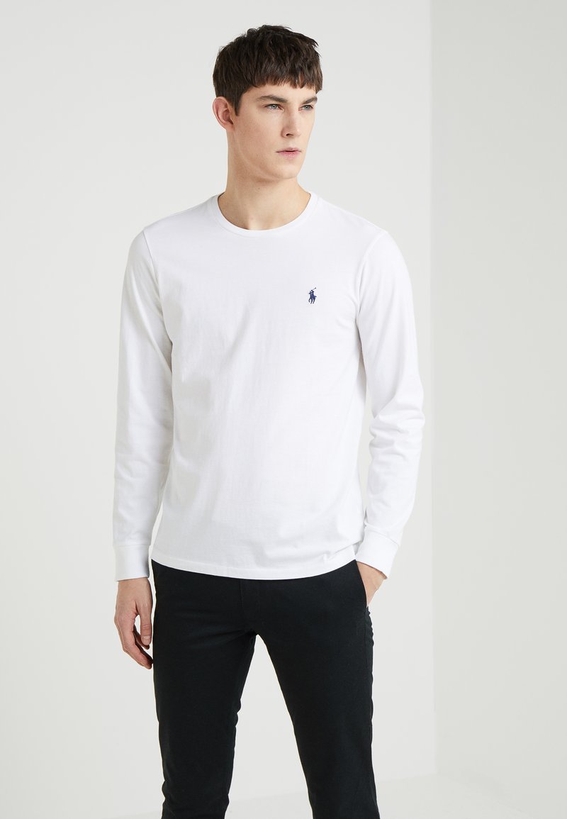 Fehér, hosszú ujjú pamut póló, kerek nyakkal és egy kis sötétkék logóval a mellkason, fekete, slim-fit nadrággal párosítva.