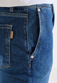 Denim jeans in medium blue with contrasting yellow stitching, featuring a side pocket, metal snap closure, and leather accent.