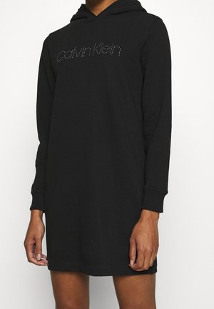 Person trägt ein schwarzes Calvin Klein Hoodie-Kleid mit langen Ärmeln und Strass-Logo auf der Brust, steht vor einem einfachen weißen Hintergrund.