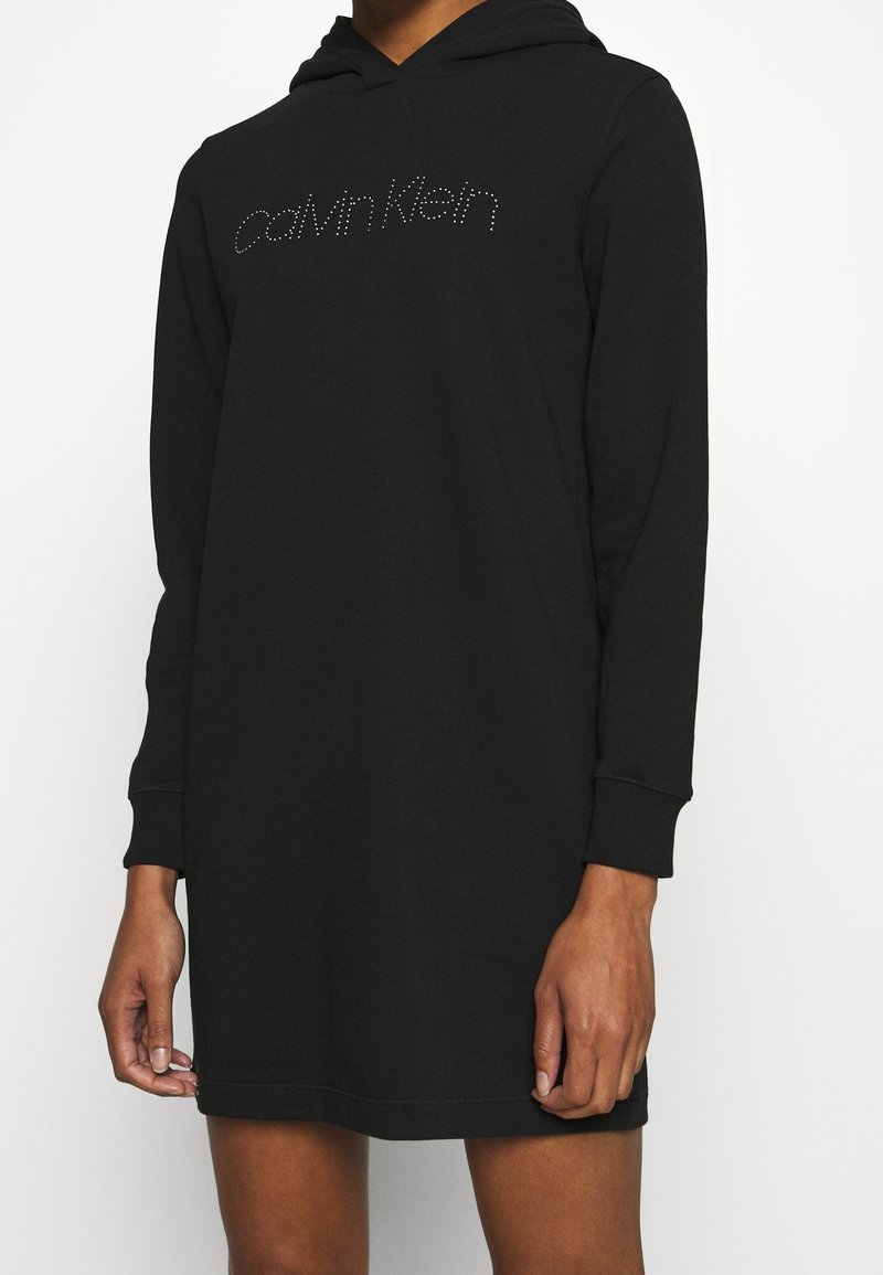 Personne portant une robe à capuche Calvin Klein noire avec manches longues et logo en strass sur la poitrine, se tenant devant un fond blanc uni.