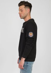 Czarna bluza z metalicznym napisem, okrągłym dekoltem, ściągaczami przy rękawach oraz wyszywanym patch'em w kształcie tygrysa na правym rękawie.