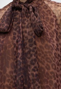 Blouse marron transparente avec imprimé léopard foncé et un grand nœud noué à l'encolure.