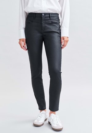 Personne portant un pantalon en cuir noir slim, une chemise blanche boutonnée et des baskets blanches, debout sur un sol gris clair.