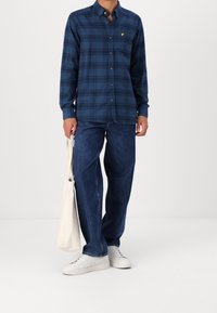 Niebieska koszula w kratę z długim rękawem i kołnierzykiem na guziki, z małym żółtym logo. Do tego luźne niebieskie dżinsy i białe trampki.
