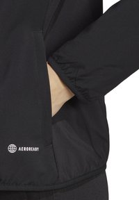 Giacca nera con design aderente, caratterizzata da una superficie texturizzata e maniche elasticizzate. Dettaglio del logo "AEROREADY" sull'orlo inferiore.