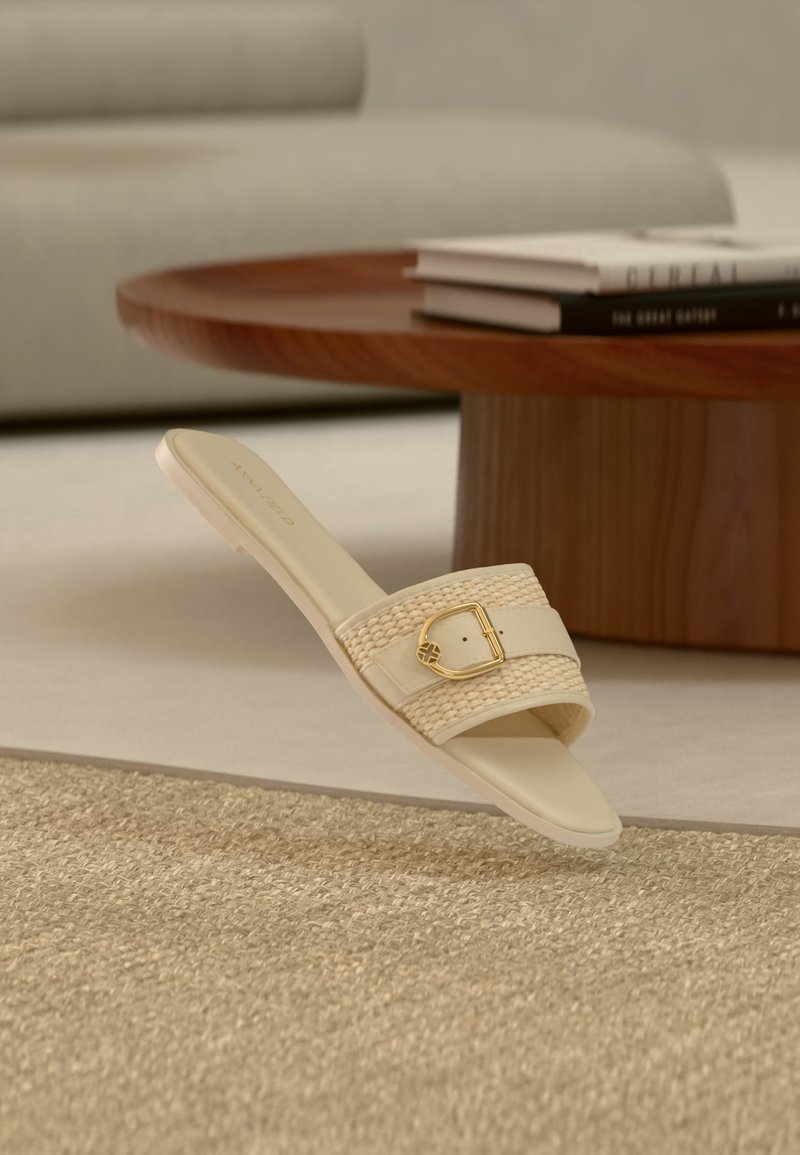 Μπεζ σανδάλι slip-on με υφαντό λουράκι και χρυσή αγκράφα, τοποθετημένο κοντά σε ξύλινο τραπεζάκι σαλονιού με βιβλία σε ένα σαλόνι ουδέτερων τόνων.