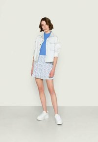 Casaco de ganga branco sobre um topo de malha azul, combinado com uma saia floral azul clara e ténis brancos. O conjunto tem um aspeto descontraído e em camadas.