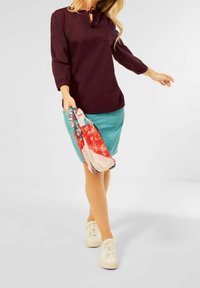 Femme marchant, portant une chemise bordeaux à manches longues, une jupe bleu sarcelle jusqu'aux genoux, des baskets blanches, tenant une écharpe rouge et bleue à motifs.