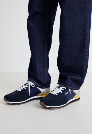 Sneakers blu navy con lacci bianchi, dettagli grigi, particolare giallo sulla suola e una tomaia in tessuto liscio, abbinati a pantaloni in denim scuro.