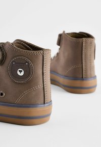 Braune High-Top-Schuhe aus strukturiertem Material, mit einem Bärenpatch an der Seite und einer Gummisohle mit blauen und braunen Streifen.