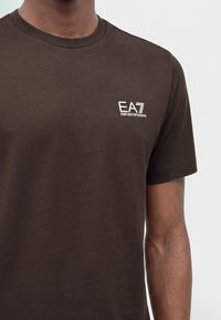 Άνδρας φοράει σκούρο καφέ κοντομάνικο μπλουζάκι με μικρό λευκό λογότυπο EA7 Emporio Armani στο αριστερό στήθος.