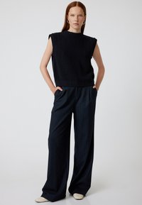Svart stickad ärmlös topp med ribbad textur, i kombination med navyrandiga vida byxor. Modellen står med händerna i fickorna och bär neutrala skor.