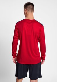 Rød langærmet træningsskjorte med marineblå skulderdetaljer, lavet af åndbart stof. Båret med sorte shorts. Glat tekstur, tætsiddende design.
