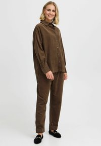 Camisa de pana marrón y pantalones a juego con un corte relajado, que cuentan con un cierre delantero abotonado, mangas largas y puños doblados. Zapatos negros sin cordones.