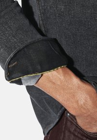 Schwarze Jeansjacke mit hochgerolltem Ärmel, ausgestattet mit kontrastierenden Nähten in Gelb und Grau, die eine strukturierte Oberfläche und sichtbare Ärmeldetails zeigt.
