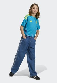 Ljus turkos tröja med gula detaljer och Adidas-logotyp, kombinerad med löst sittande blå denimjeans med sidostråkar och svarta skor.