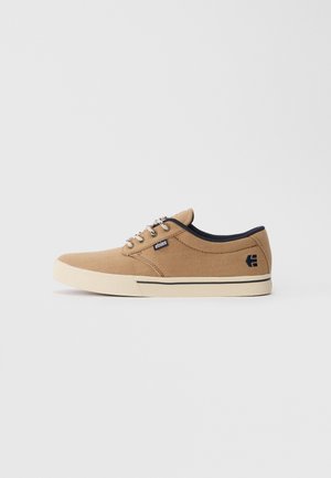 Sneaker in tela beige a taglio basso con suola bianca, bordi neri, lacci con motivo e logo "etnies" nero sul lato e sul tallone.