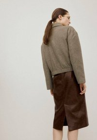 Kobieta z brązowym kucykiem, nosząca okulary, brązową kratowaną kurtkę oraz ciemnobrązową skórzaną spódnicę midi z tylnego rozcięcia, stojąca na jednolitym tle.