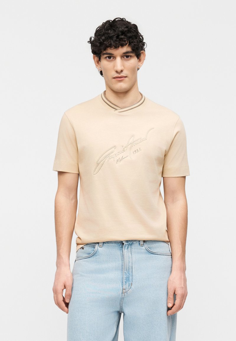 Νεαρός άνδρας με σγουρά μαλλιά, φορώντας μπεζ κο ντομάνικο πουκάμισο με κείμενο σε γραφή και ανοιχτόχρωμο μπλε τζιν, στέκεται μπροστά σε απλό φόντο.