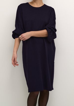 Personne portant une robe bleu marine foncé mi-longue avec des manches longues bouffantes et un ourlet côtelé, associée à des collants noirs transparents et une bague argentée.
