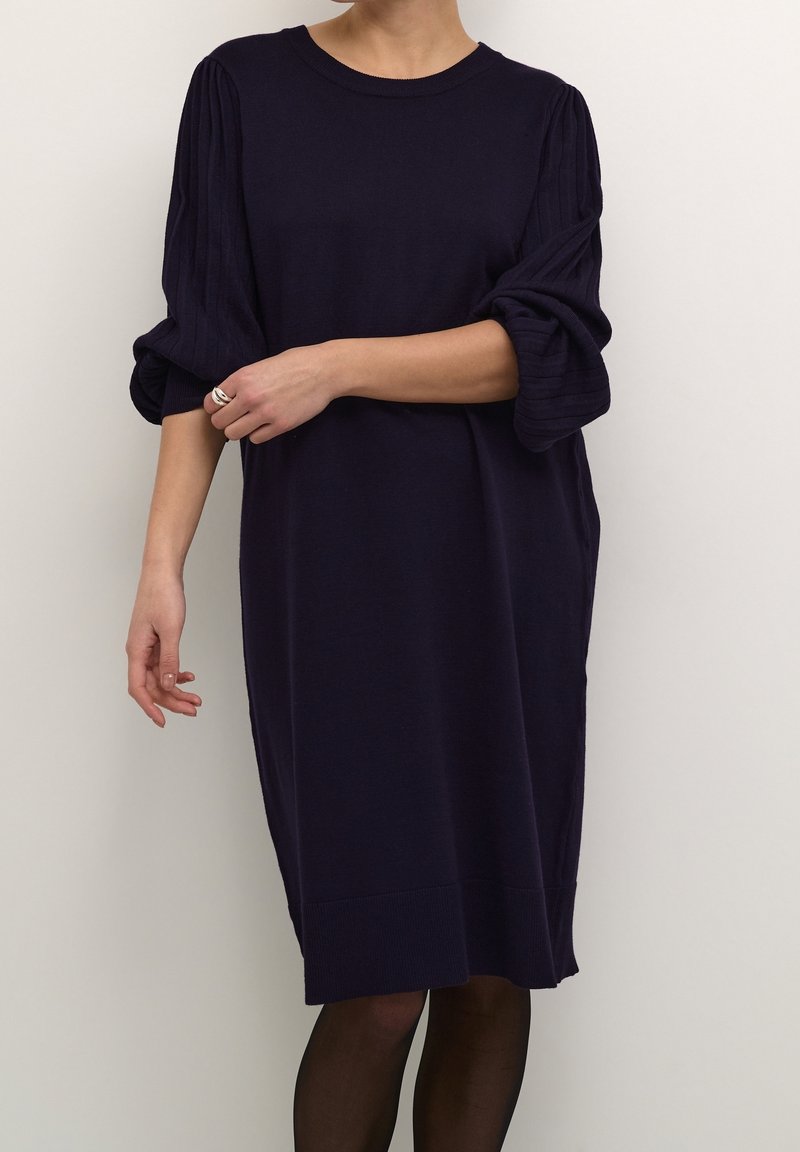Personne portant une robe bleu marine foncé mi-longue avec des manches longues bouffantes et un ourlet côtelé, associée à des collants noirs transparents et une bague argentée.