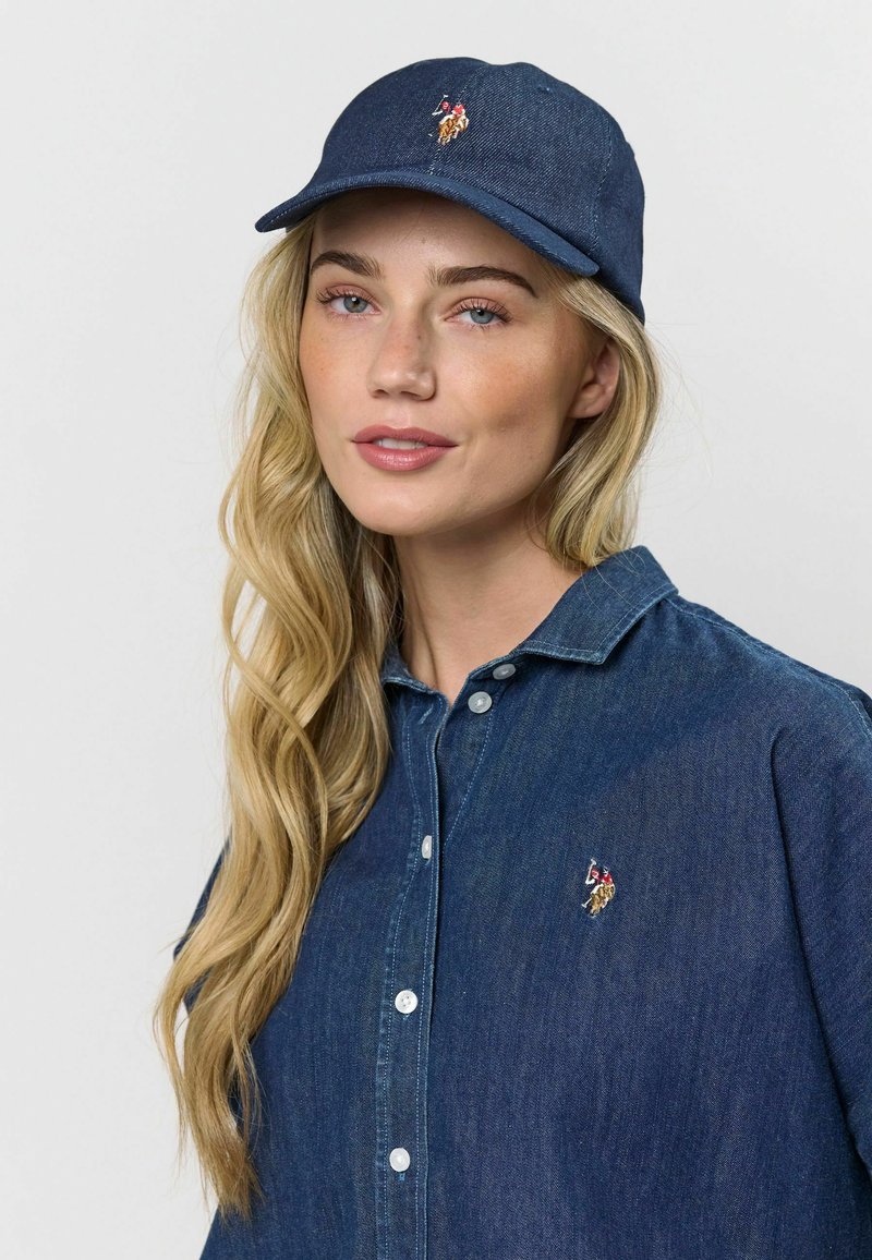 Tmavomodrá denimová čiapka s ohnutým šiltom a vyšitým logom; v kombinácii s tmavomodrou denimovou košeľou na gombíky s viditeľným šitím a gombíkmi.