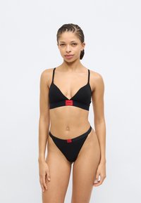 Black bra and thong set featuring a triangular design, thin straps, and a red logo band on both pieces, made from smooth fabric.