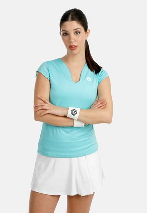 Femme aux cheveux bruns attachés en queue de cheval, portant un haut à manches courtes bleu clair, une jupe de tennis blanche et des bracelets blancs, debout les bras croisés.