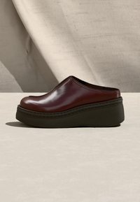 Σκούρο κόκκινο δερμάτινο παπούτσι χωρίς κορδόνια με παχύ μαύρο σόλα πλατφόρμας, τοποθετημένο σε ανοιχτή επιφάνεια με μπεζ ύφασμα στο φόντο.