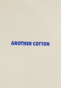 Krém színű pamut anyag, élénk kék hímzéssel, amelyen a "ANOTHER COTTON" felirat található. Egyszerű, letisztult dizájn, sima tapintással.