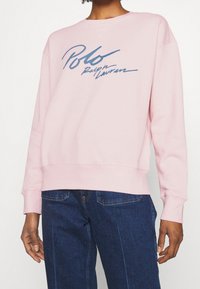 Ljusrosa sweatshirt med rund halsringning och långa ärmar. Den har en marinblå logotypdesign på framsidan. Mjukt bomullsmaterial.