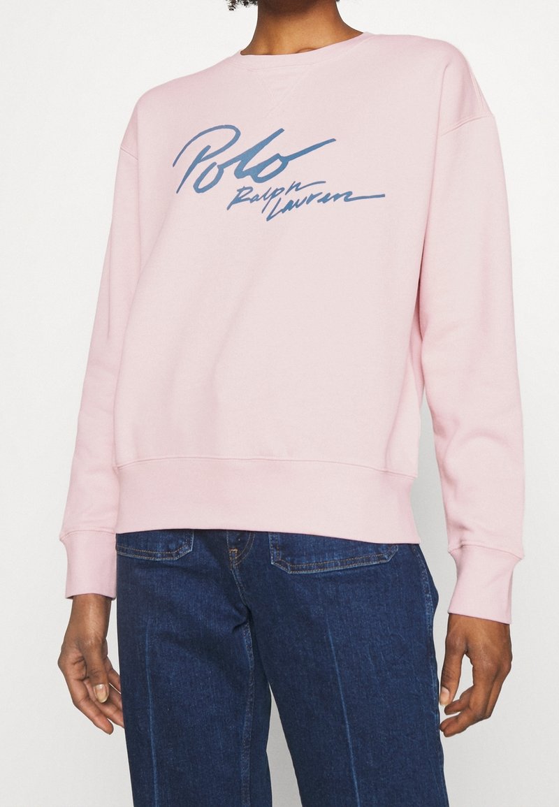 Ljusrosa sweatshirt med rund halsringning och långa ärmar. Den har en marinblå logotypdesign på framsidan. Mjukt bomullsmaterial.