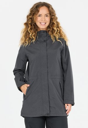 Lächelnde Frau mit lockigem Haar, die eine dunkelgraue lange Jacke mit Fronttaschen trägt und vor einem schlichten weißen Hintergrund steht.