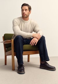 Ljusbeige texturerad tröja, mörka jeans och mörka snörskor med robusta sulor, sittande på en grön stoppad trästol.