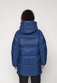 Marineblauwe gewatteerde jas met een capuchon, voorzien van horizontale gestikte secties, een langere achterzoom en elastische manchetten voor comfort.