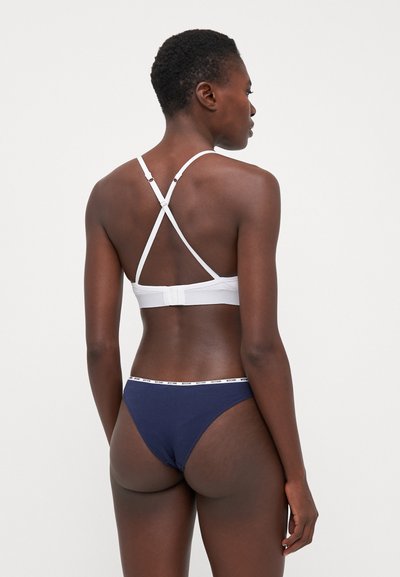 White bralette with crisscross straps and back closure; paired with navy blue thong featuring a white branded waistband.