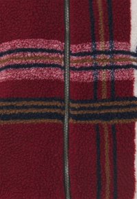 Veste polaire rouge et rose à surface texturée. Présente des rayures à carreaux horizontales en marron, marine et bleu clair, avec une fermeture éclair argentée.