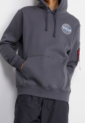 Osoba w ciemnoszarej bluzie NASA z kieszenią z przodu i kieszenią na rękawie z zamkiem, ręka w kieszonce z przodu, na jasnym, jednolitym tle.