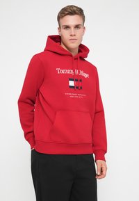 Red hooded sweatshirt made of cotton blend, featuring a front pocket, embroidered logo, and graphic design with blue, white, and red accents.