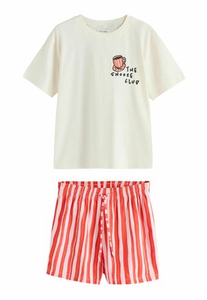 Jonge vrouw met lang bruin haar draagt een wit T-shirt met een afbeelding van een koffiekopje en rode gestreepte korte broek, staand met handen in de zakken.